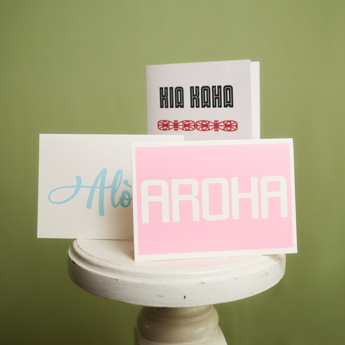 Three greeting cards displayed on a stand against a green background, with the words 'KIA KAHA', 'Alofa', and 'AROHA' respectively, showcased Te Reo and Pasifika cards