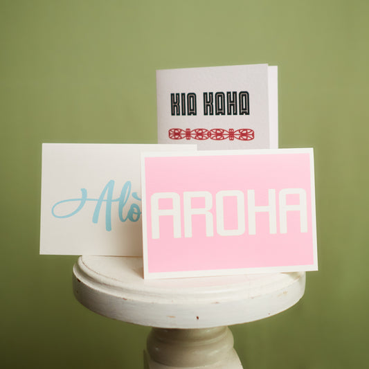 Three greeting cards displayed on a stand against a green background, with the words 'KIA KAHA', 'Alofa', and 'AROHA' respectively, showcased Te Reo and Pasifika cards